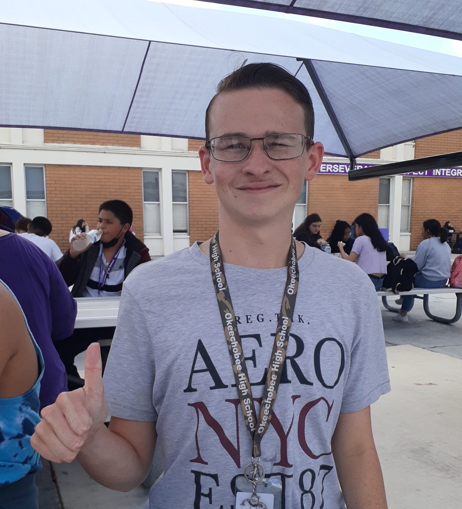 It's a boy smiling and giving a thumbs-up, he is wearing a grey shirt with text on it. Along with a camo lanyard, and thin, black, rectangle glasses.