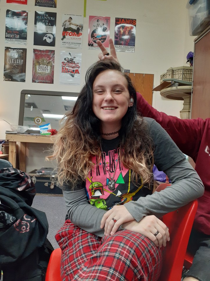 It's a picture of a high school girl sitting in a red chair. She has her hair down, which is black that fades into blond. In this picture, she is wearing a black t-shirt over a grey long sleeve shirt, and red pants.