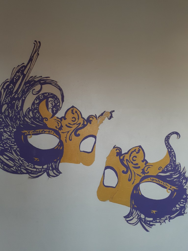 It shows a painting of two domino masks, there both looking towards each other and the mask has gold and purple. On the purple side it looks like feathers are on the mask and the other side is just a smooth gold with some purple designs.