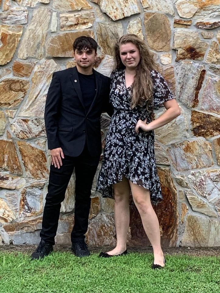 Grace Thomas stands on the right in a white on black floral dress while Colton stands on the left in a black suit.