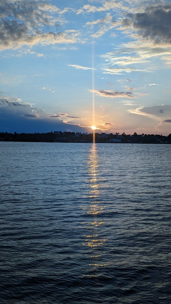 The sun is shining beautifully on the water and it creates a line of sunlight that reflects and makes the water glisten. The sky is a beautiful shade of blue mixed with a purple hue and the clouds are all scattered while the sun sits in the middle.