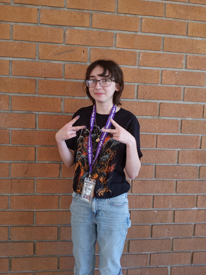 Bright is doing the peace sign on both hands. She is wearing a black T-shirt with some kind of graphics on it and she is wearing light blue jeans. She has on her school ID and she has short black hair. She has on black reading glasses and has in no earrings.